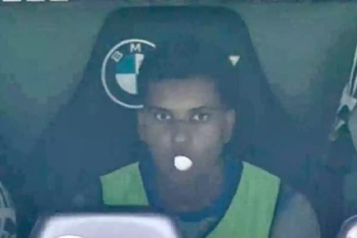 Person sitting on a sports bench wearing a fluorescent green vest and a white pill in their mouth Person sitting on a sports bench wearing a fluorescent green vest and a white pill in their mouth