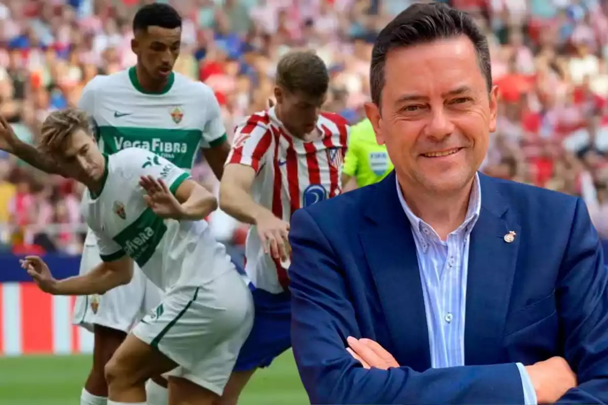 Un hombre de traje sonríe al frente mientras al fondo se juega un partido de fútbol entre jugadores con uniformes blanco y verde y rojo con blanco.