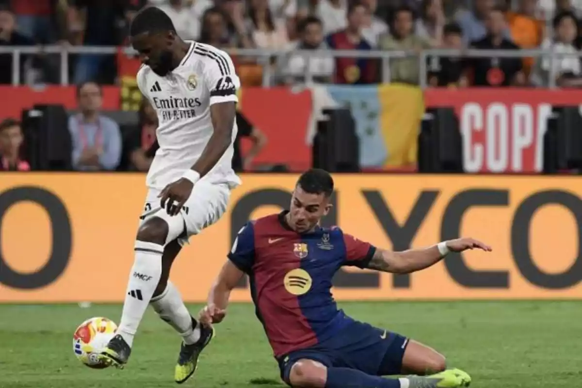 Un jugador del Real Madrid controla el balón mientras un jugador del Barcelona intenta quitarle la pelota deslizándose en el césped durante un partido de fútbol