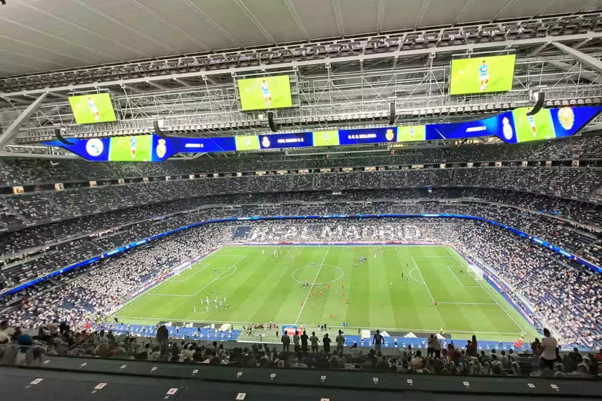 Estadio lleno de aficionados durante un partido de fútbol con pantallas gigantes y el césped iluminado Estadio lleno de aficionados durante un partido de fútbol con pantallas gigantes y el césped iluminado