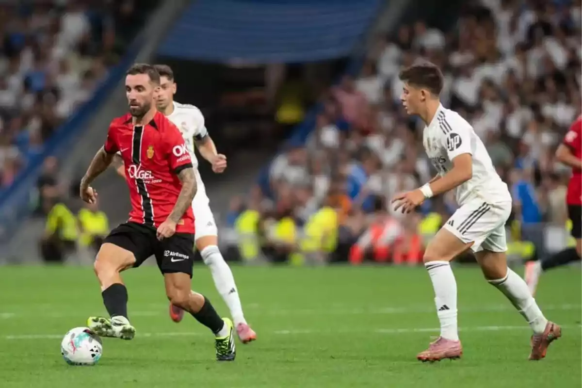 Un jugador con uniforme rojo y negro controla el balón mientras dos jugadores con uniforme blanco lo presionan durante un partido de fútbol en un estadio lleno de espectadores. Un jugador con uniforme rojo y negro controla el balón mientras dos jugadores con uniforme blanco lo presionan durante un partido de fútbol en un estadio lleno de espectadores.