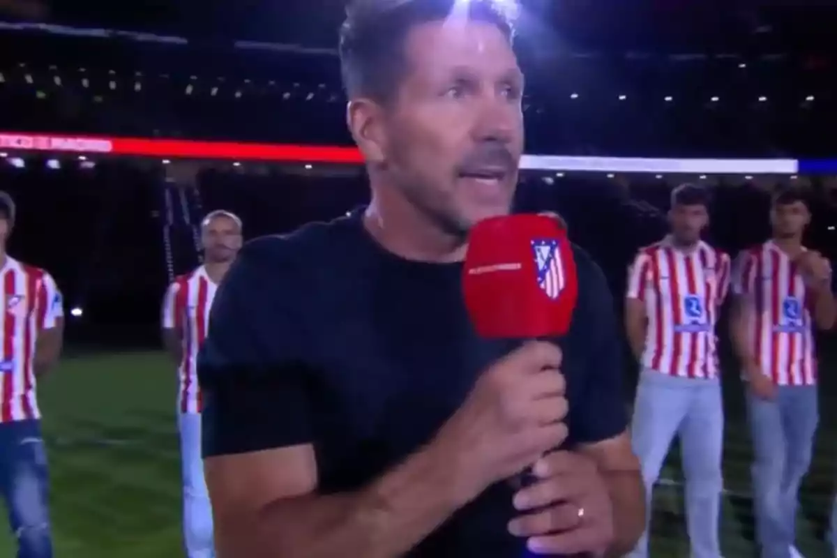 Un hombre sostiene un micrófono rojo con el escudo del Atlético de Madrid mientras habla en un estadio iluminado, al fondo se ven jugadores con uniforme de rayas rojas y blancas. Un hombre sostiene un micrófono rojo con el escudo del Atlético de Madrid mientras habla en un estadio iluminado, al fondo se ven jugadores con uniforme de rayas rojas y blancas.