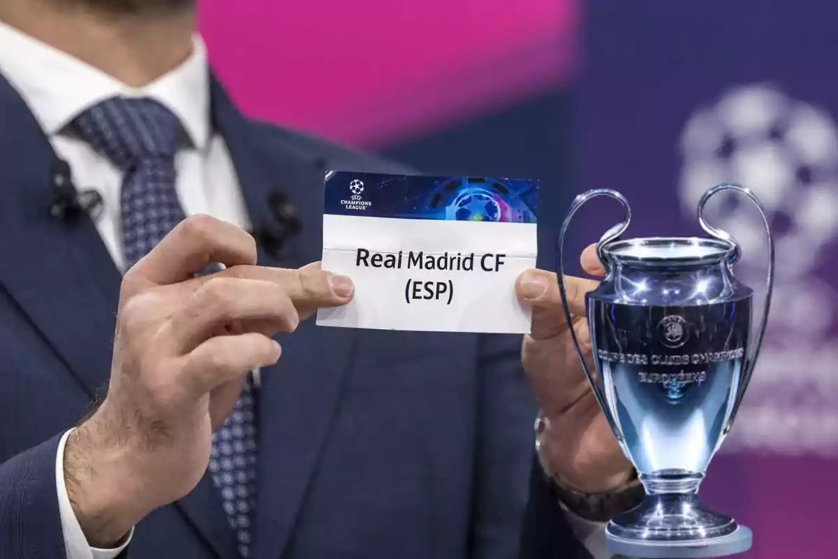 Person in a suit holding a paper with the name Real Madrid CF next to a Champions League trophy