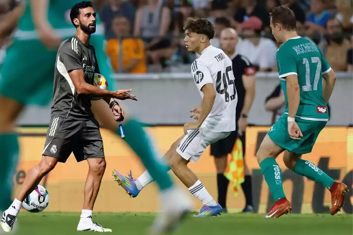 Entrenador de fútbol con uniforme del Real Madrid en la cancha mientras dos jugadores disputan el balón durante un partido