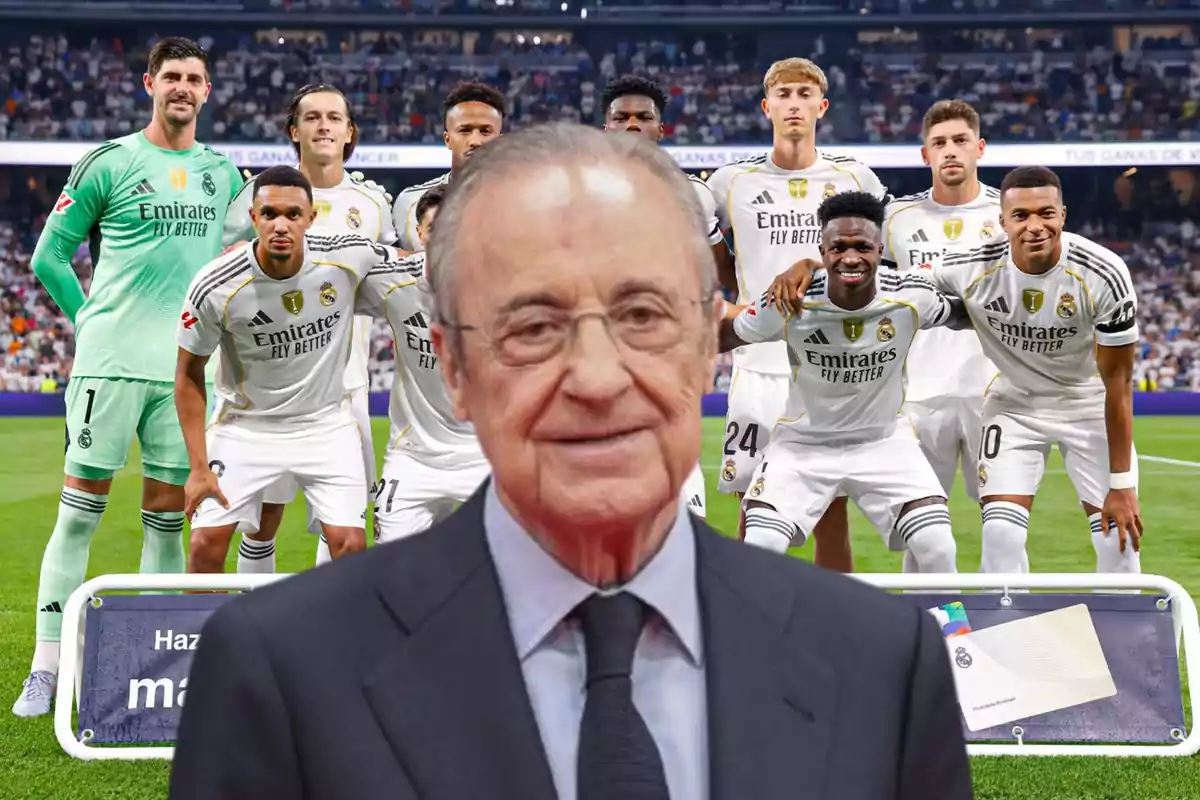 An older man wearing glasses and a suit appears in the foreground in front of a group of players in white uniforms posing in a stadium full of spectators.