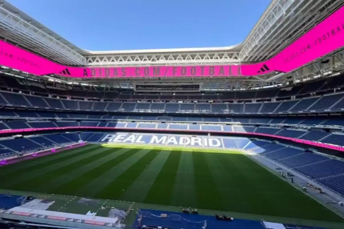 Estadio de fútbol moderno con gradas azules y el nombre Real Madrid visible en los asientos, rodeado de pantallas publicitarias rosas y césped bien cuidado bajo un cielo despejado Estadio de fútbol moderno con gradas azules y el nombre Real Madrid visible en los asientos, rodeado de pantallas publicitarias rosas y césped bien cuidado bajo un cielo despejado
