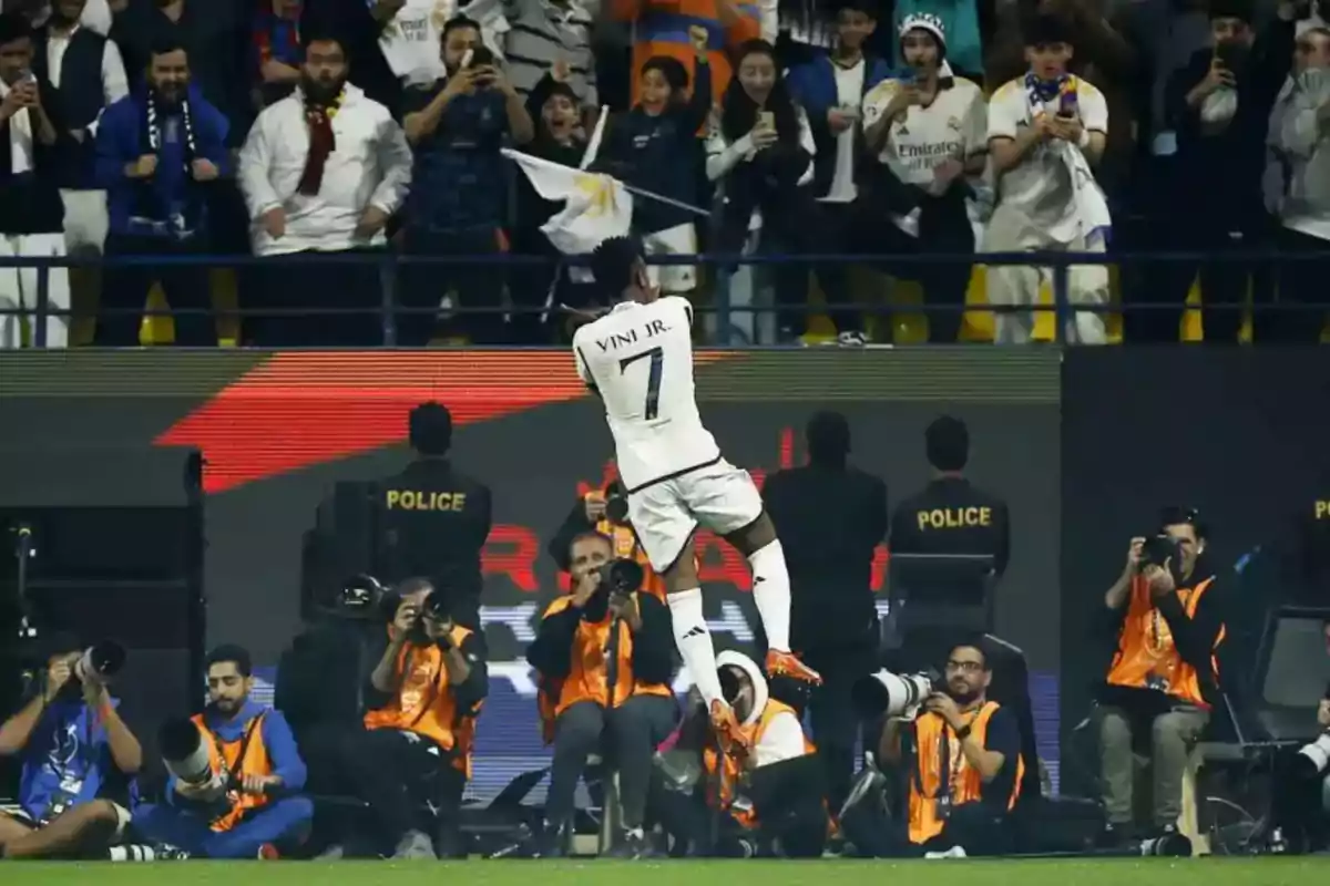 Futbolista celebrando un gol saltando frente a la afición mientras fotógrafos capturan el momento y policías vigilan el área Futbolista celebrando un gol saltando frente a la afición mientras fotógrafos capturan el momento y policías vigilan el área