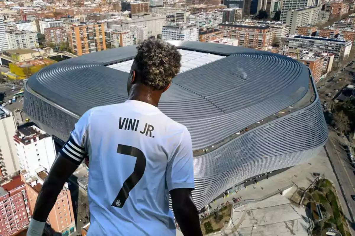 A player named Vini Jr., wearing the number 7 on his jersey, appears in the foreground with his back to the viewer, facing a large, modern stadium in a city.