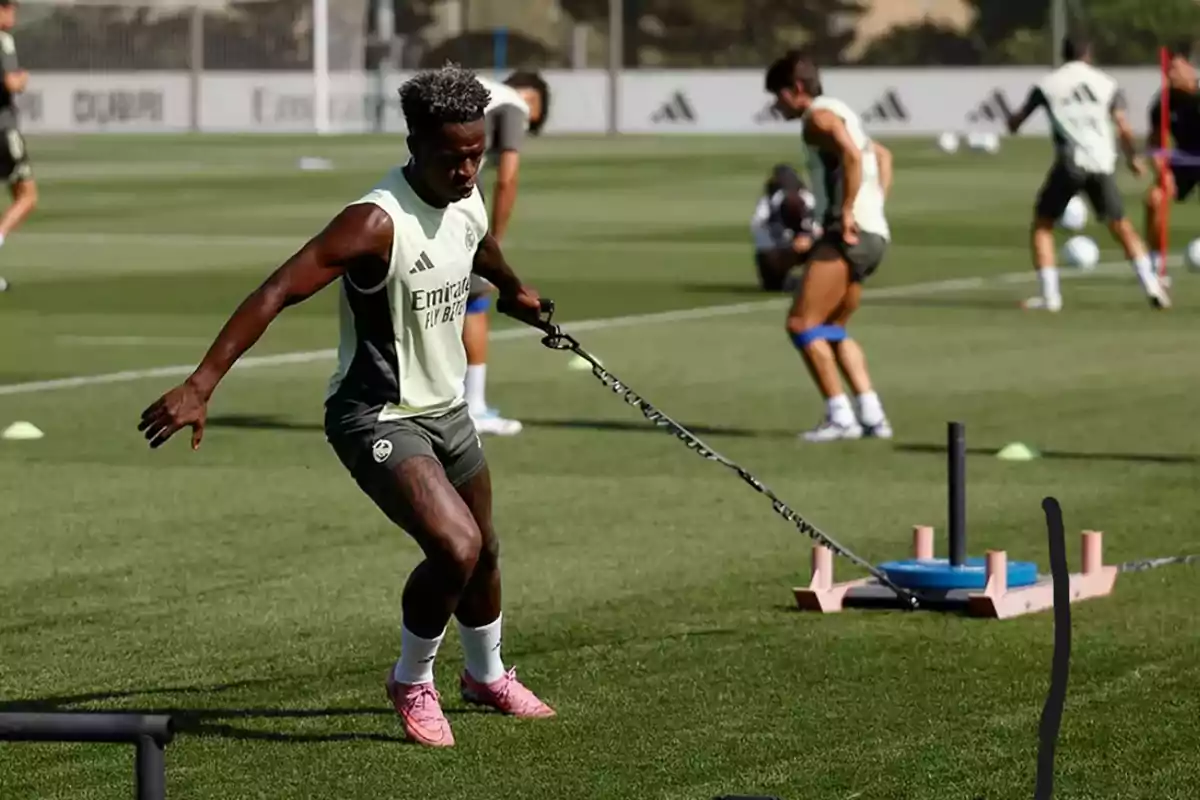 Soccer player training on a grass field while dragging a weight sled with a chain, with other players practicing in the background.