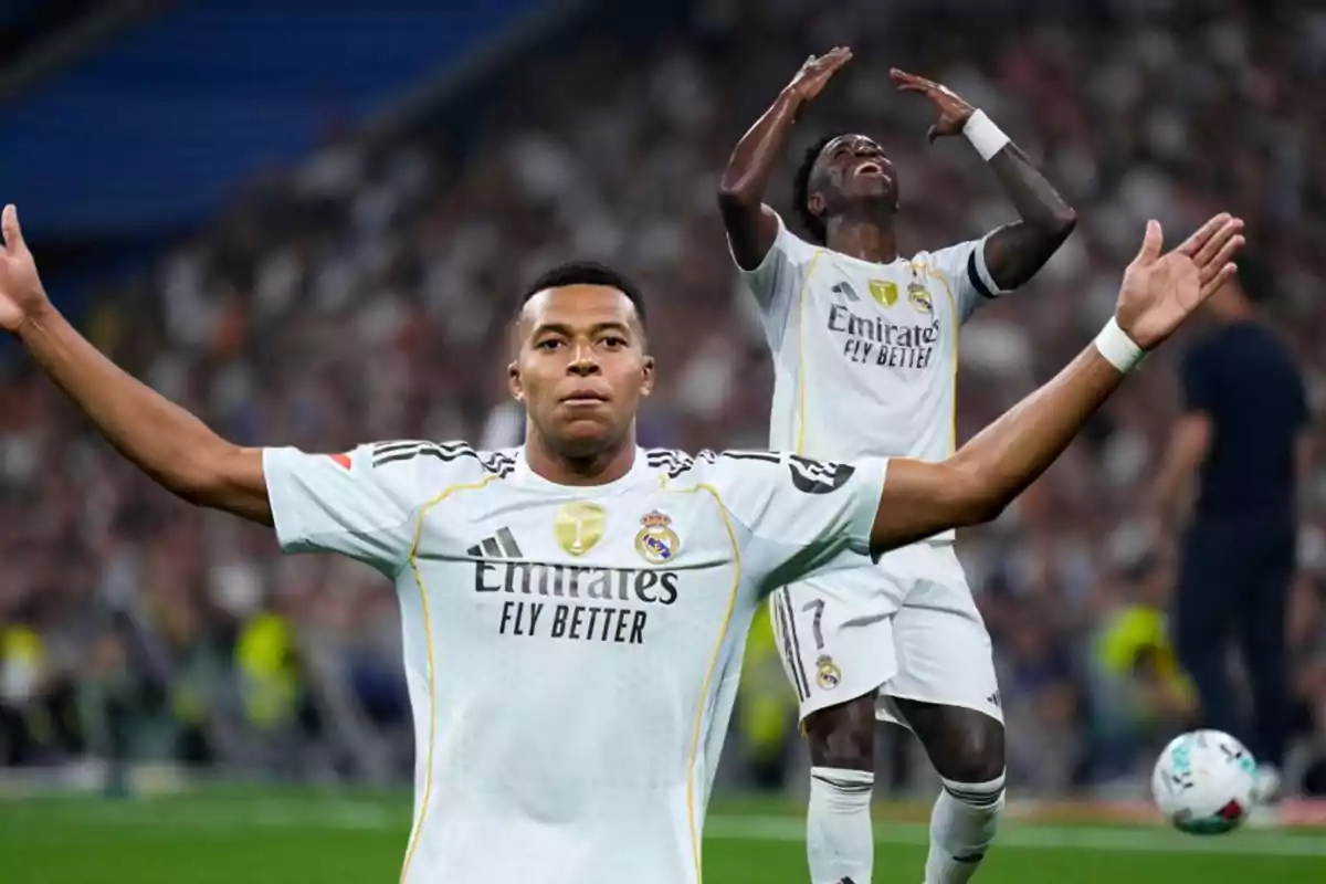 Dos futbolistas con uniforme blanco del Real Madrid celebran en el campo durante un partido, uno con los brazos extendidos y otro levantando las manos hacia el cielo.