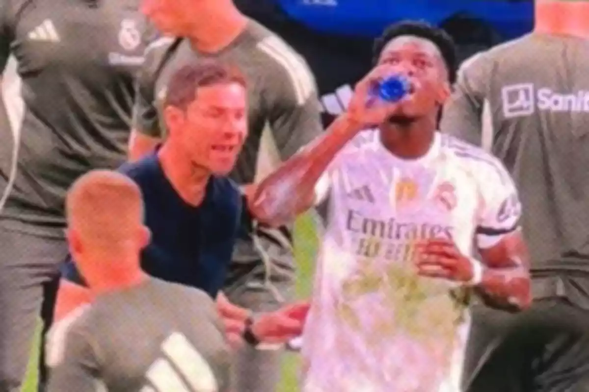 A player in a white uniform drinks water while several people in sportswear surround him on the field.