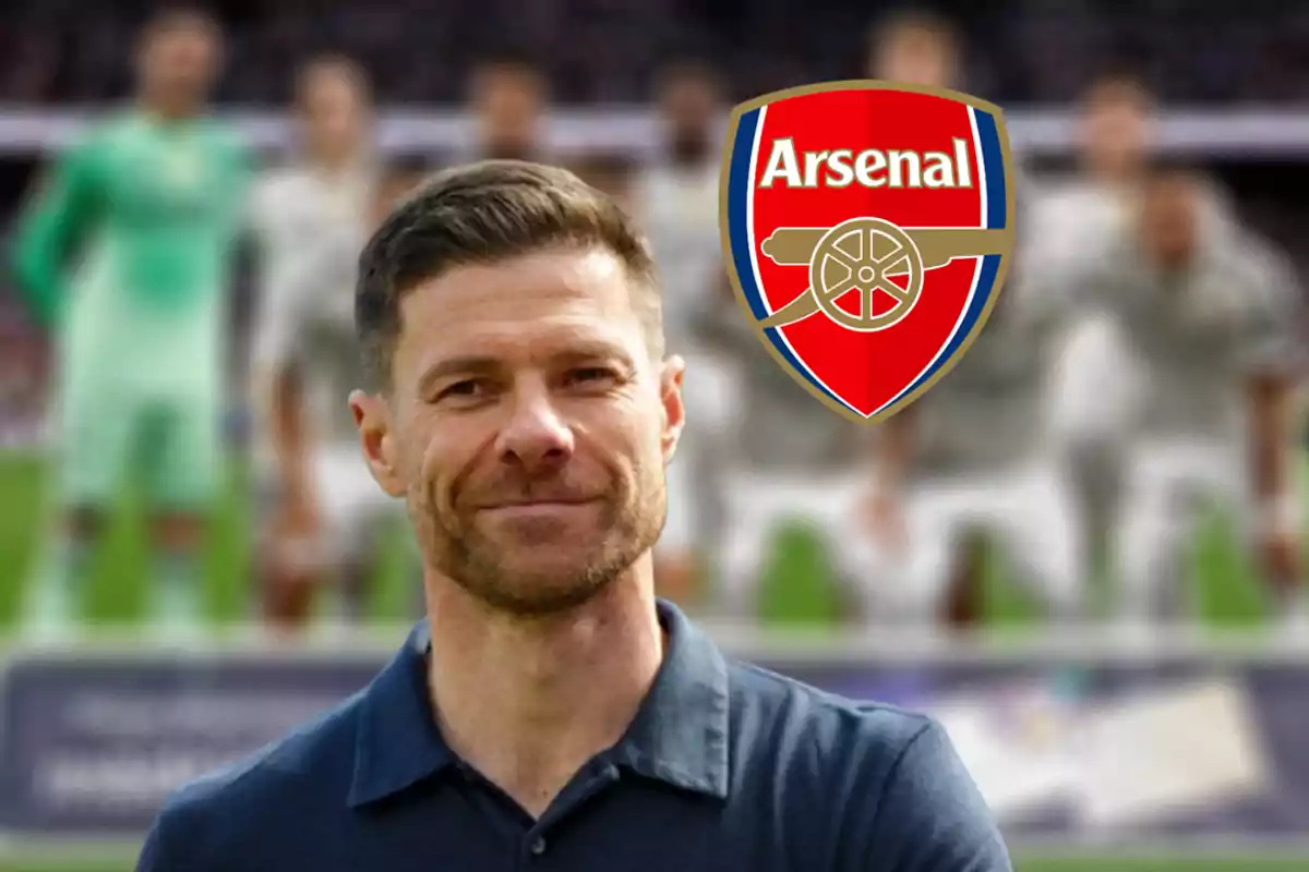 Short-haired, bearded man wearing a dark blue t-jersey smiles in front of the Arsenal crest with a blurred soccer team in the background.
