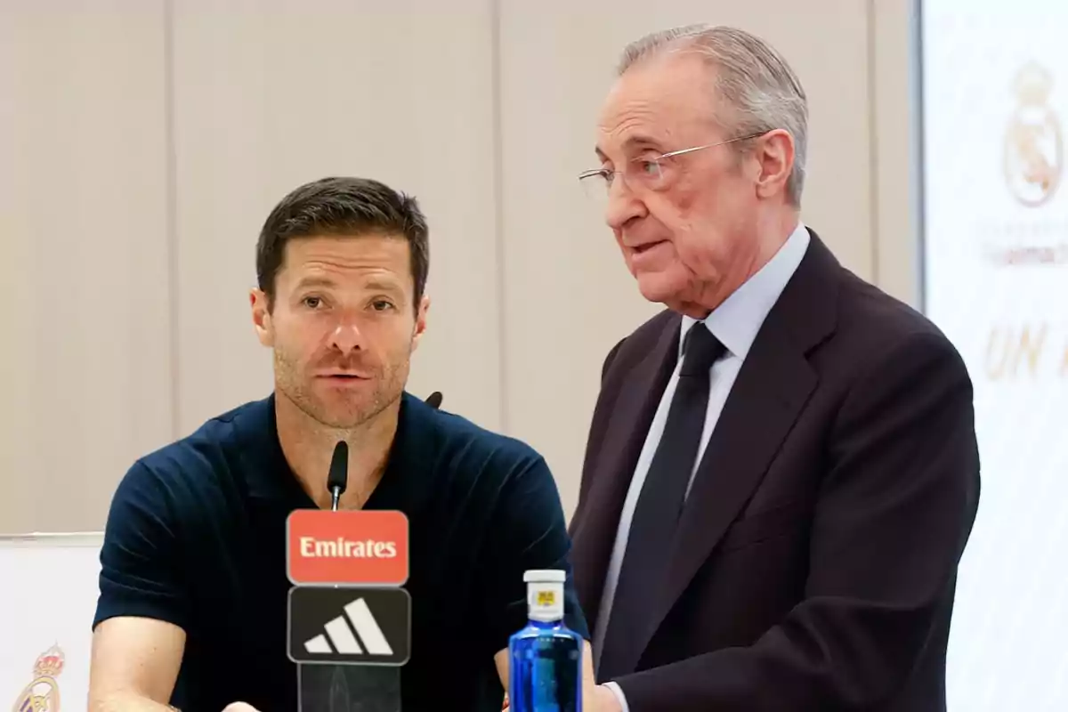 Two men at a press conference with a microphone and a bottle of water on the table; one of them is wearing a suit and the other is dressed in a dark polo jersey.