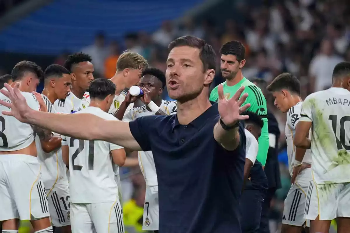 A soccer coach gestures with open arms in front of a group of players dressed in white uniforms on a field.
