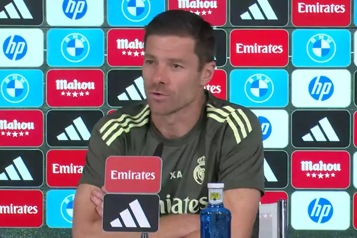 Entrenador del Real Madrid sentado en una rueda de prensa con fondo de patrocinadores y una botella de agua sobre la mesa