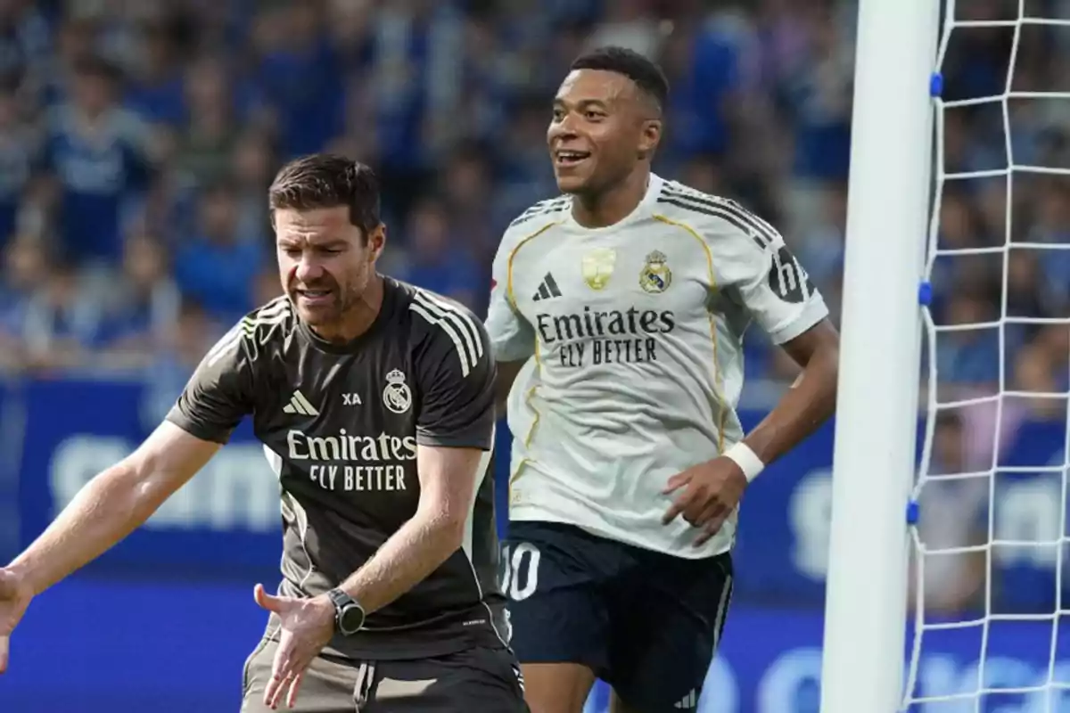 Dos personas con uniforme del Real Madrid en un estadio de fútbol una de ellas parece estar dando indicaciones y la otra sonríe cerca de la portería