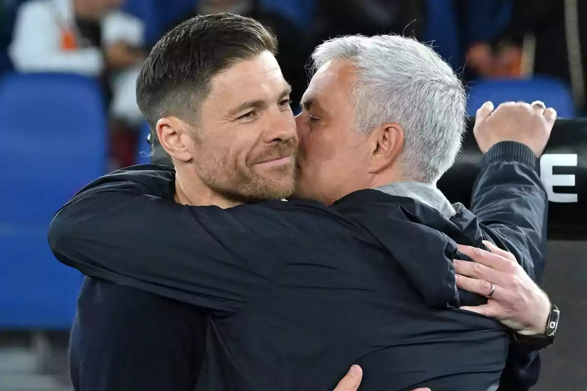 Two men hug in a soccer stadium, one of them kisses the other on the cheek while both smile slightly.