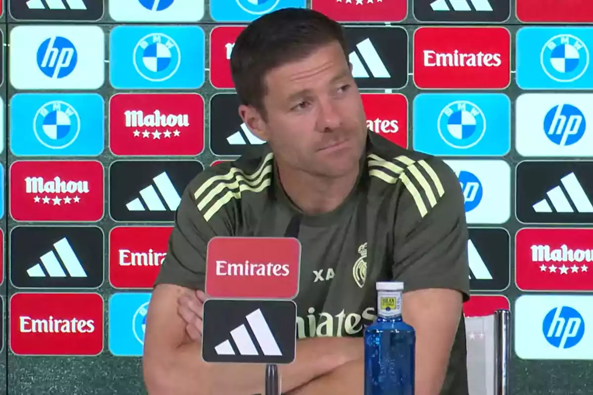 Entrenador de fútbol sentado en una rueda de prensa con fondo de patrocinadores y micrófono frente a él