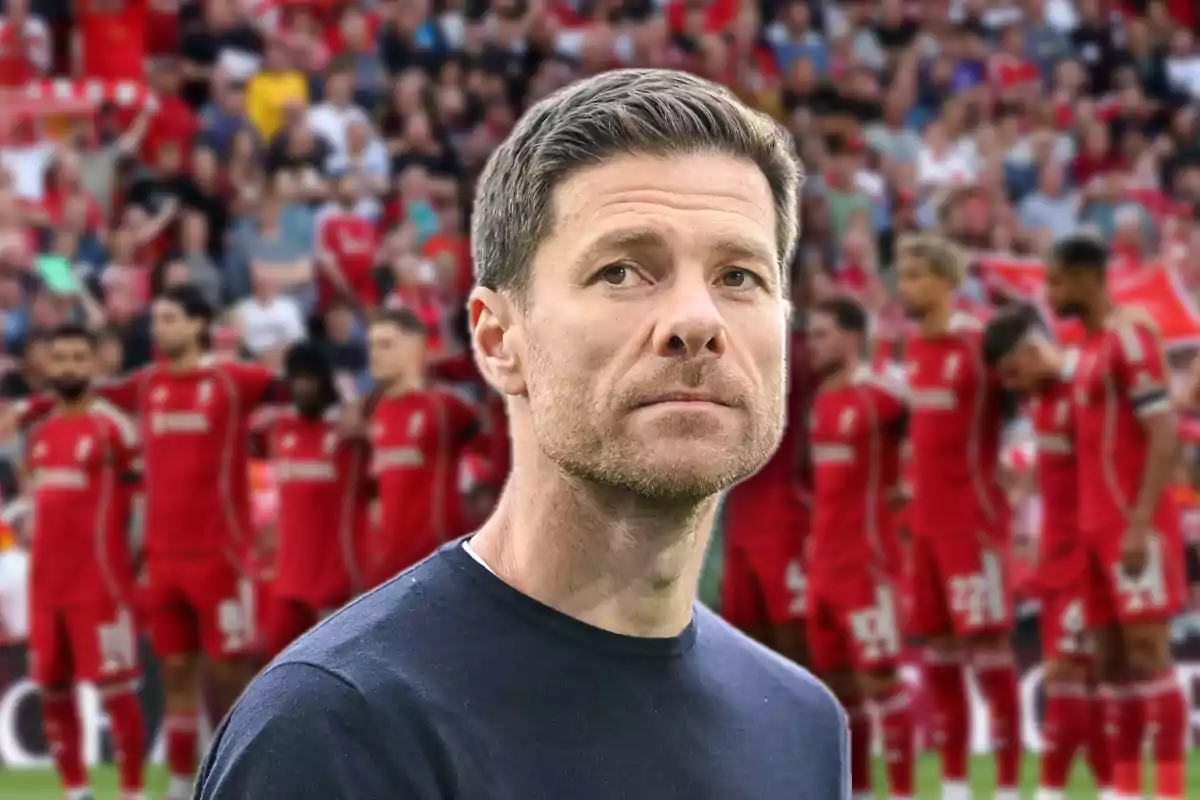 Short-haired man with a light beard and a serious expression standing in front of a group of players dressed in red in a stadium full of fans.