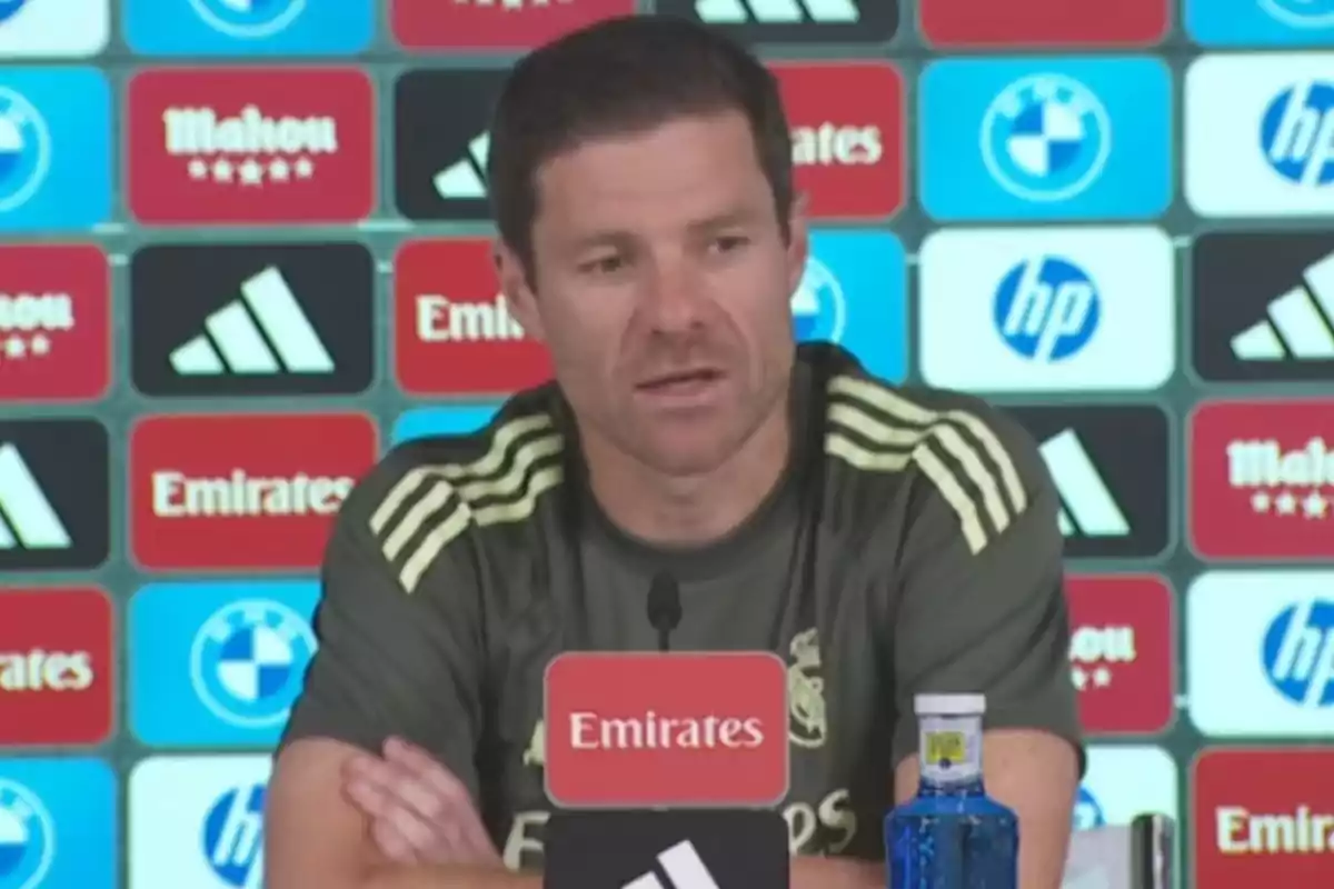 Un hombre con camiseta deportiva del Real Madrid habla en una rueda de prensa con un micrófono frente a él y un fondo lleno de logotipos de patrocinadores.