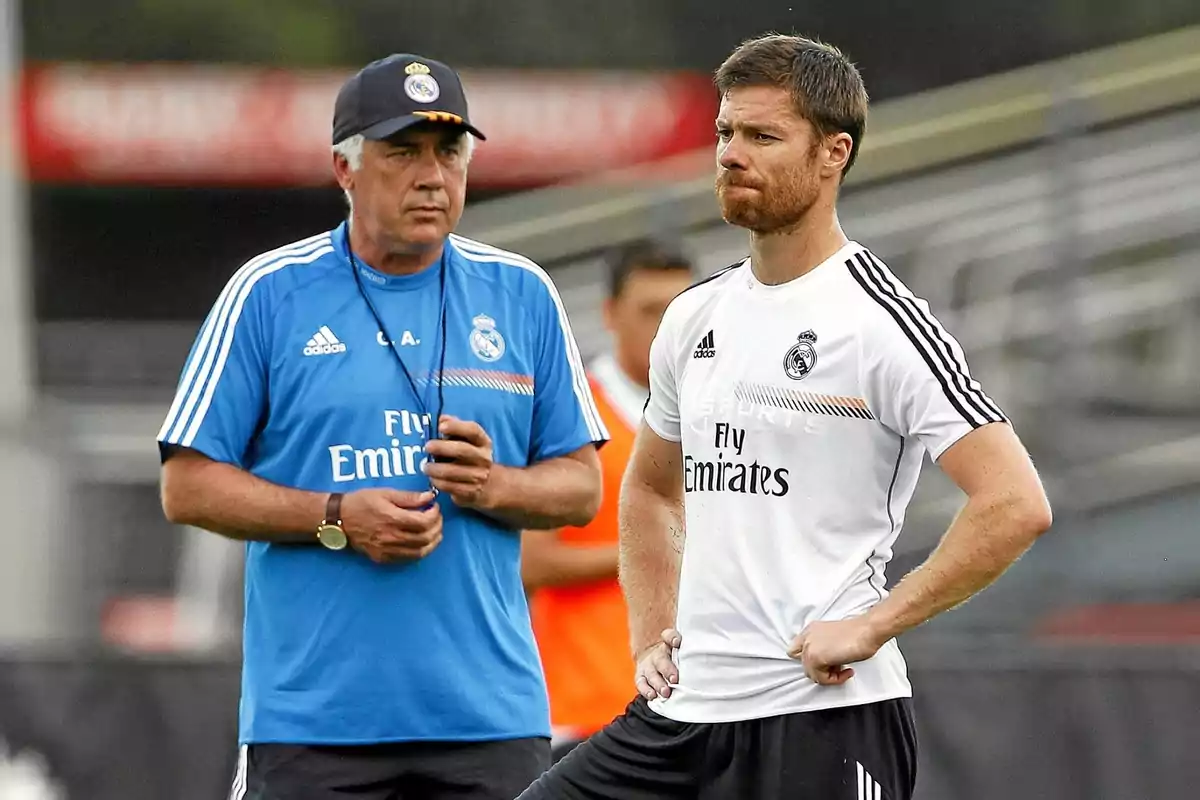 Two people wearing Real Madrid training gear are on a soccer field; one is wearing a blue jersey and a cap, the other a white jersey, and both seem focused on the conversation or on observing the training session. Two people wearing Real Madrid training gear are on a soccer field; one is wearing a blue jersey and a cap, the other a white jersey, and both seem focused on the conversation or on observing the training session.