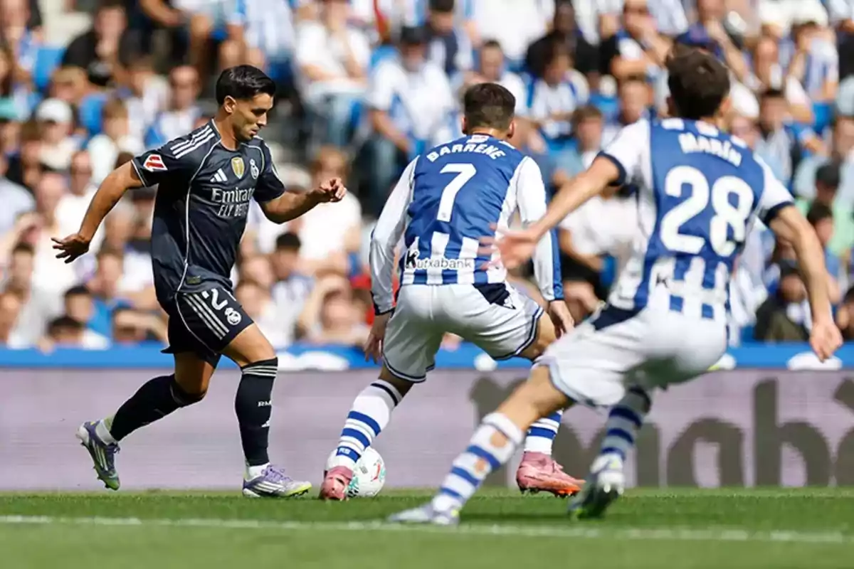 A Real Madrid player advances with the ball while two Real Sociedad players try to stop him during a soccer match in a stadium full of fans. A Real Madrid player advances with the ball while two Real Sociedad players try to stop him during a soccer match in a stadium full of fans.