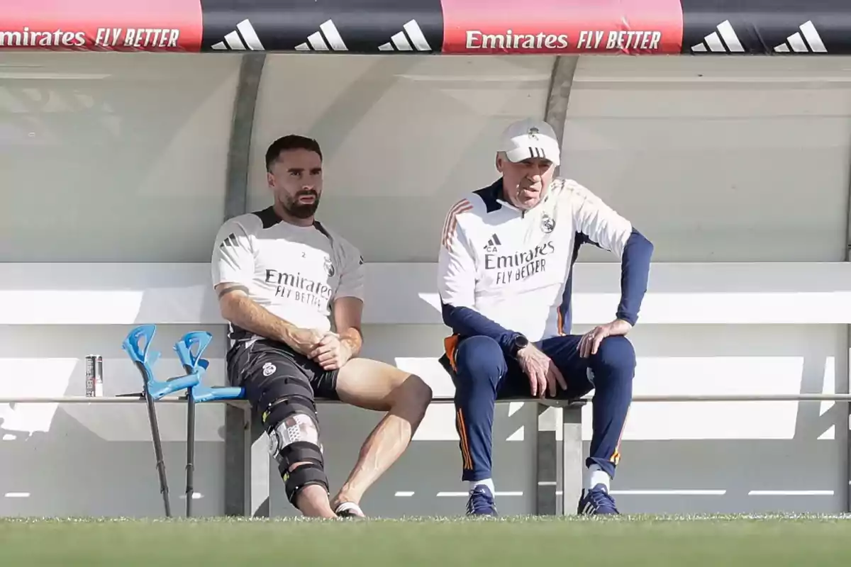 Two people wearing Real Madrid training gear are sitting on a bench; one of them has a bandaged leg and crutches resting beside them. Two people wearing Real Madrid training gear are sitting on a bench; one of them has a bandaged leg and crutches resting beside them.