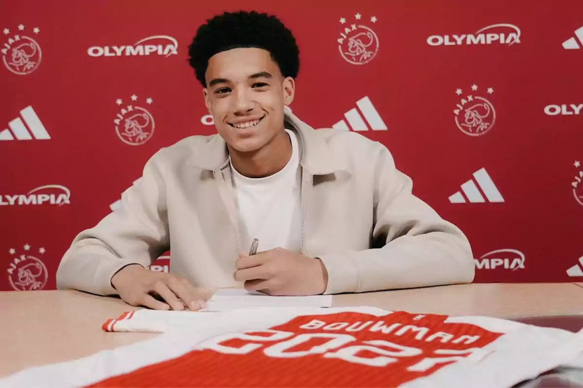 Smiling young man signing a contract with an Ajax jersey on the table and a red background with club logos Smiling young man signing a contract with an Ajax jersey on the table and a red background with club logos
