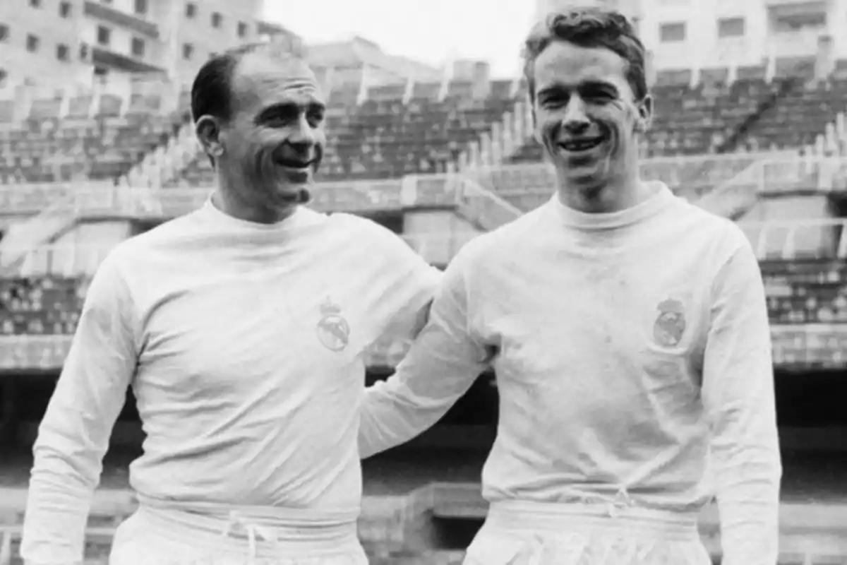 Two players in white Real Madrid uniforms pose smiling in an old stadium. Two players in white Real Madrid uniforms pose smiling in an old stadium.