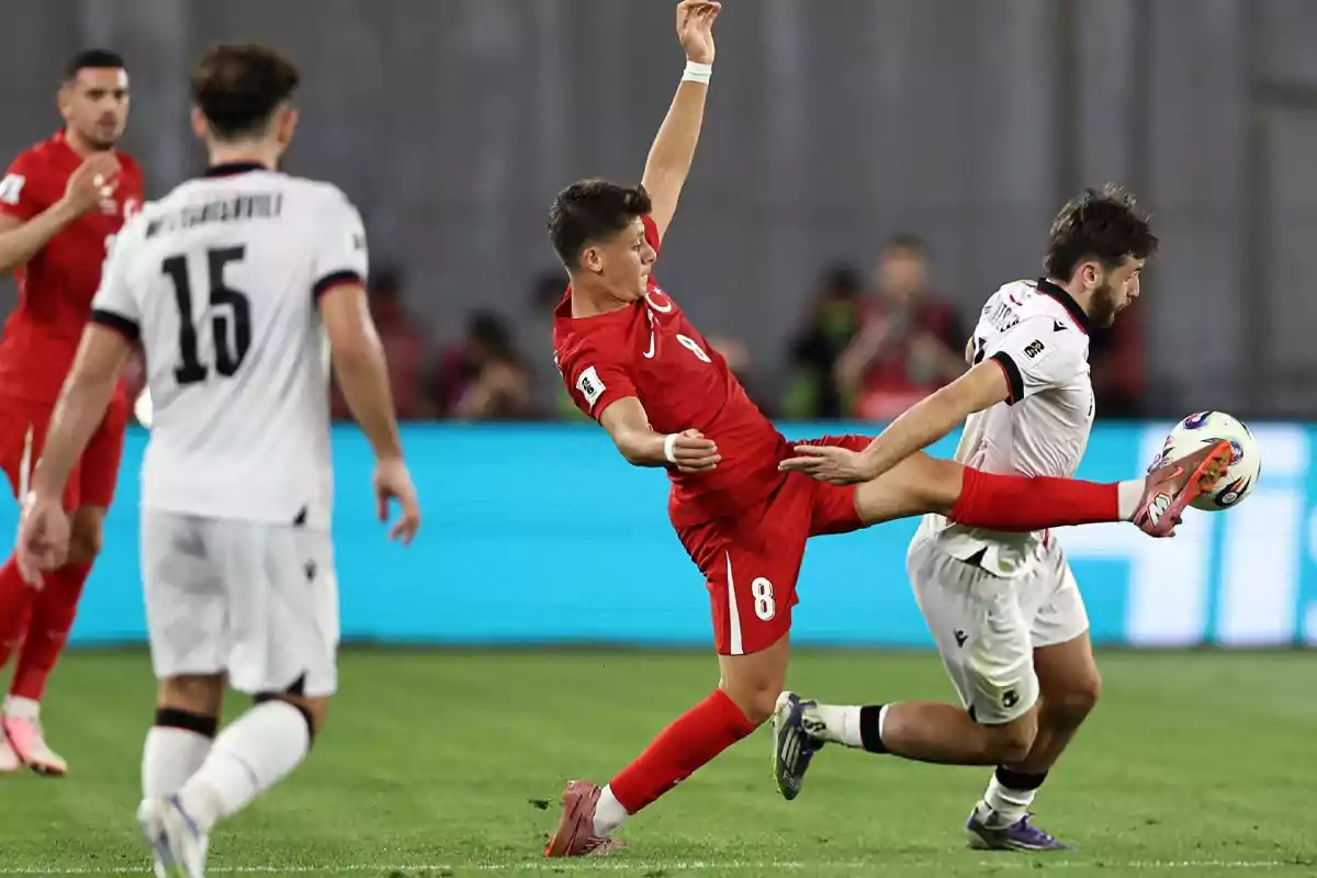 Two players compete for the ball in an intense play during a match, one dressed in red and the other in white, while other players watch the action on the field. Two players compete for the ball in an intense play during a match, one dressed in red and the other in white, while other players watch the action on the field.