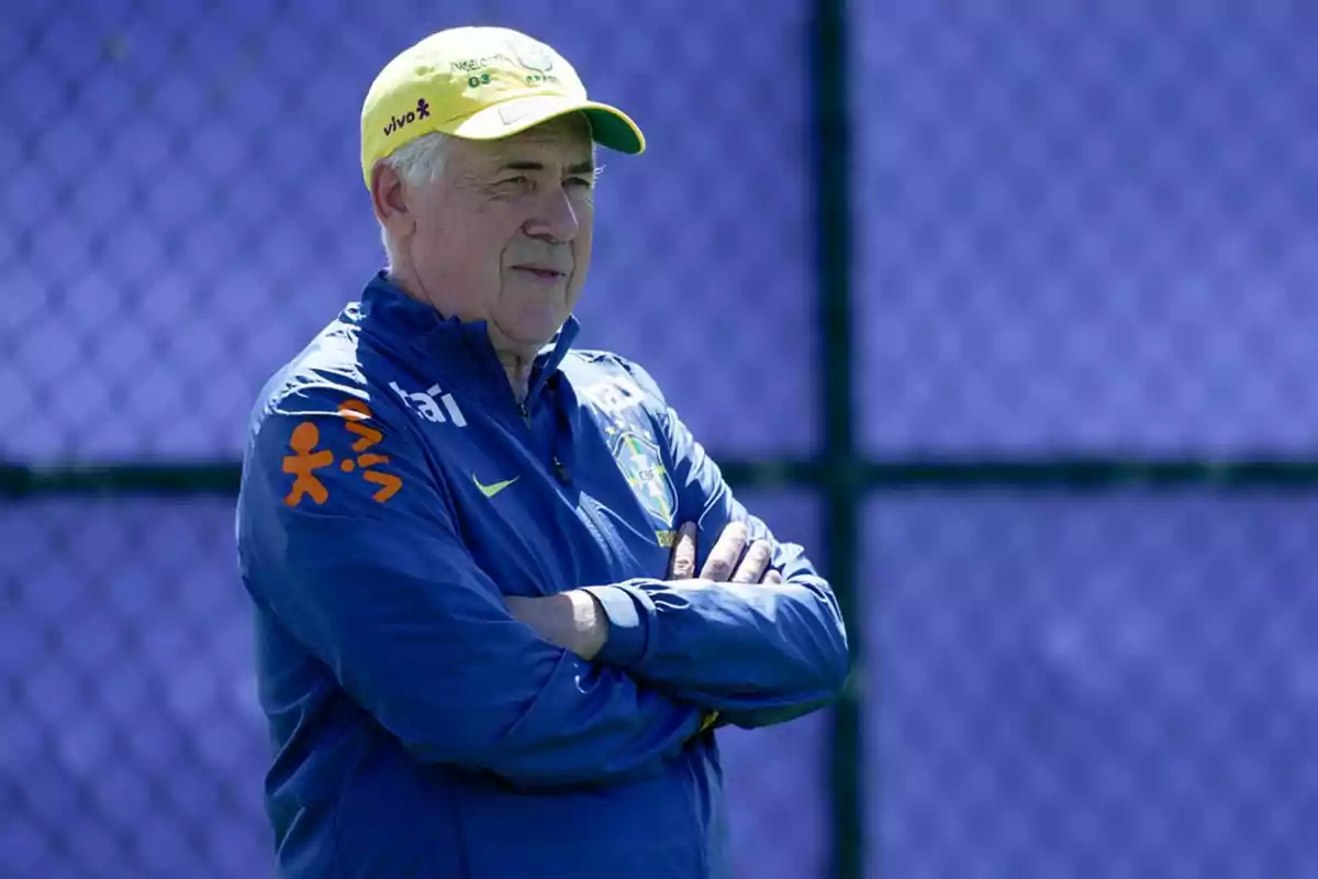 Soccer coach wearing a blue jacket and a yellow cap watching the field with arms crossed in front of a blue fence Soccer coach wearing a blue jacket and a yellow cap watching the field with arms crossed in front of a blue fence