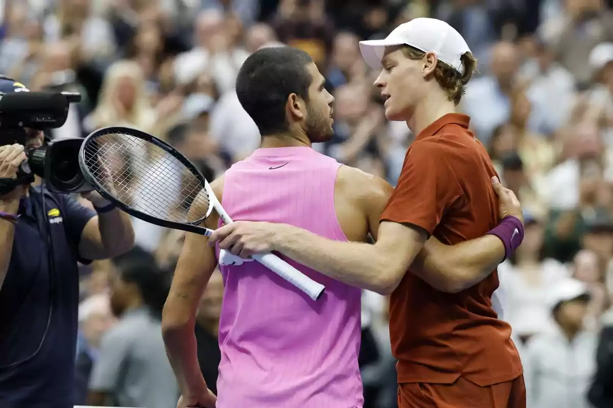 Two tennis players hug at the end of a match while a camera records them and the crowd watches in the background. Two tennis players hug at the end of a match while a camera records them and the crowd watches in the background.