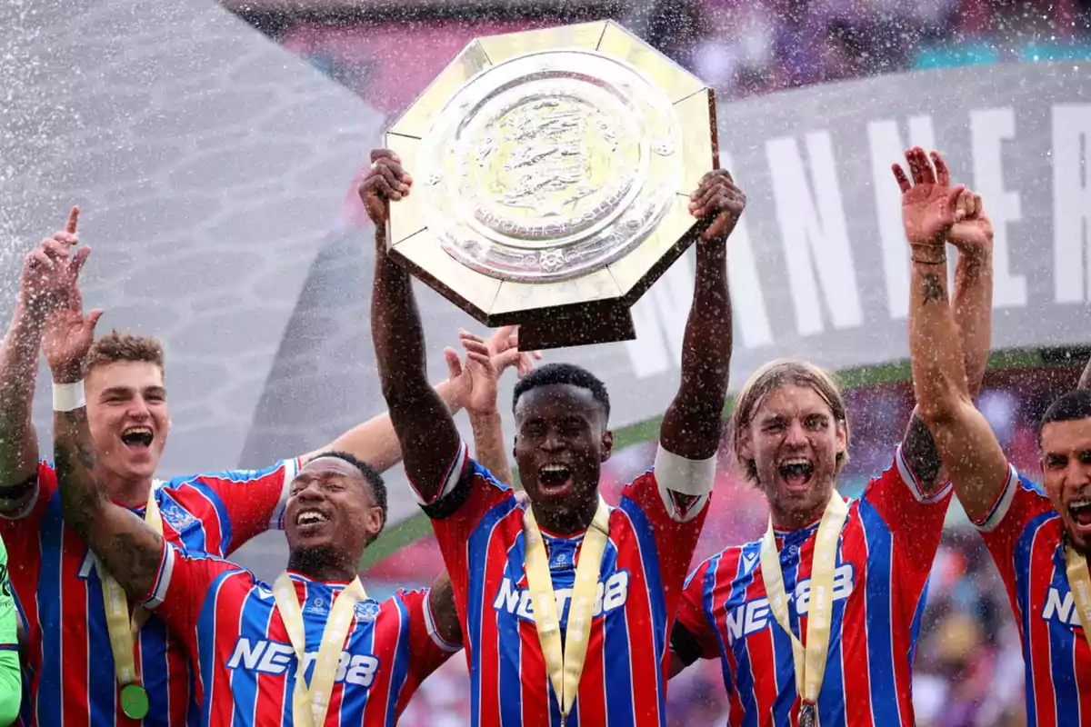 Jugadores de fútbol celebran levantando un trofeo grande y octagonal mientras son rociados con agua, todos visten uniformes a rayas rojas y azules y muestran medallas doradas Jugadores de fútbol celebran levantando un trofeo grande y octagonal mientras son rociados con agua, todos visten uniformes a rayas rojas y azules y muestran medallas doradas