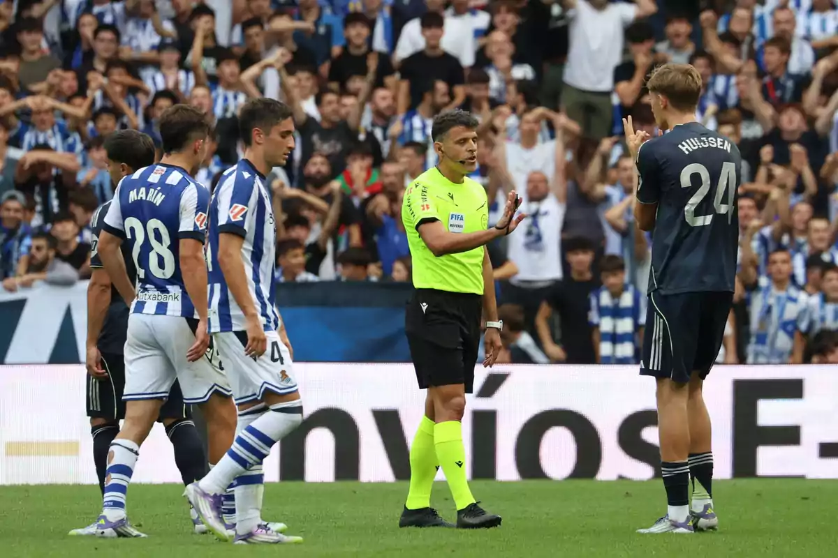 Un árbitro de fútbol detiene el juego mientras varios jugadores de dos equipos conversan en el campo y el público observa desde las gradas.