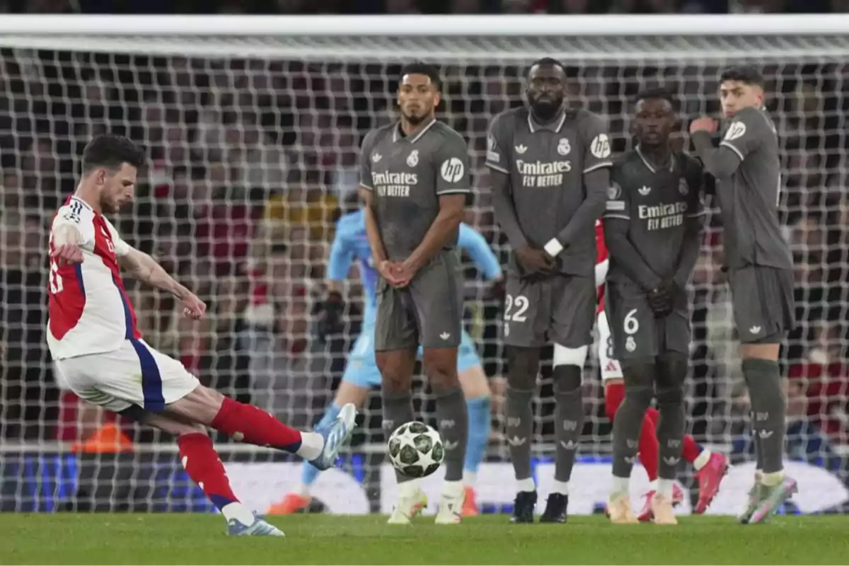 A player in a red and white uniform takes a free kick while four players in gray uniforms form a wall in front of the goal. A player in a red and white uniform takes a free kick while four players in gray uniforms form a wall in front of the goal.