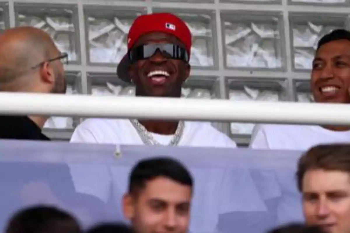 A man wearing a red cap and dark glasses smiles while sitting next to other people in the stands.