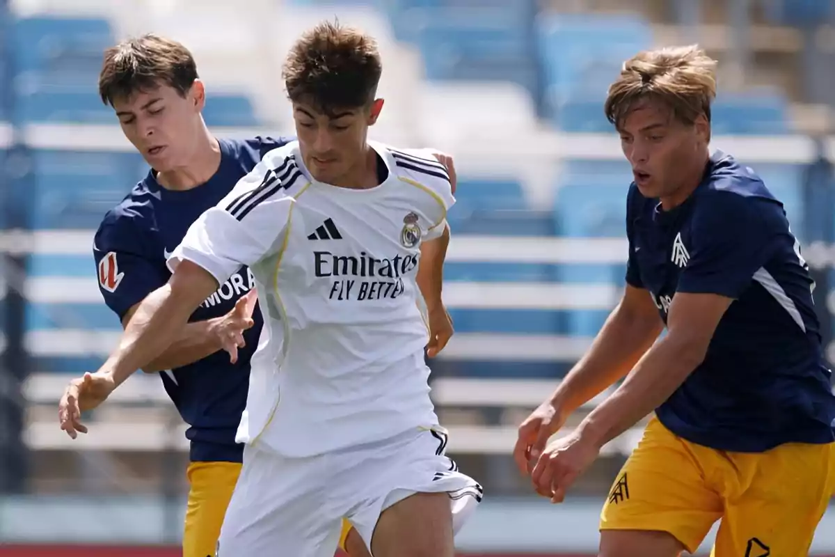 Three young players compete for the ball during a match in a stadium with blue stands; one of them is wearing a white uniform and the other two are wearing blue uniforms with yellow shorts.
