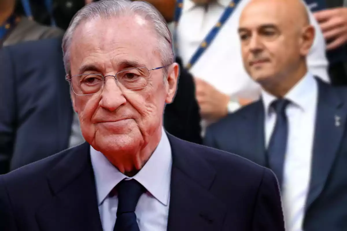 Older man with gray hair and glasses in the foreground with a serious expression, dressed in a dark suit and tie, while another bald person also in a suit appears blurred in the background