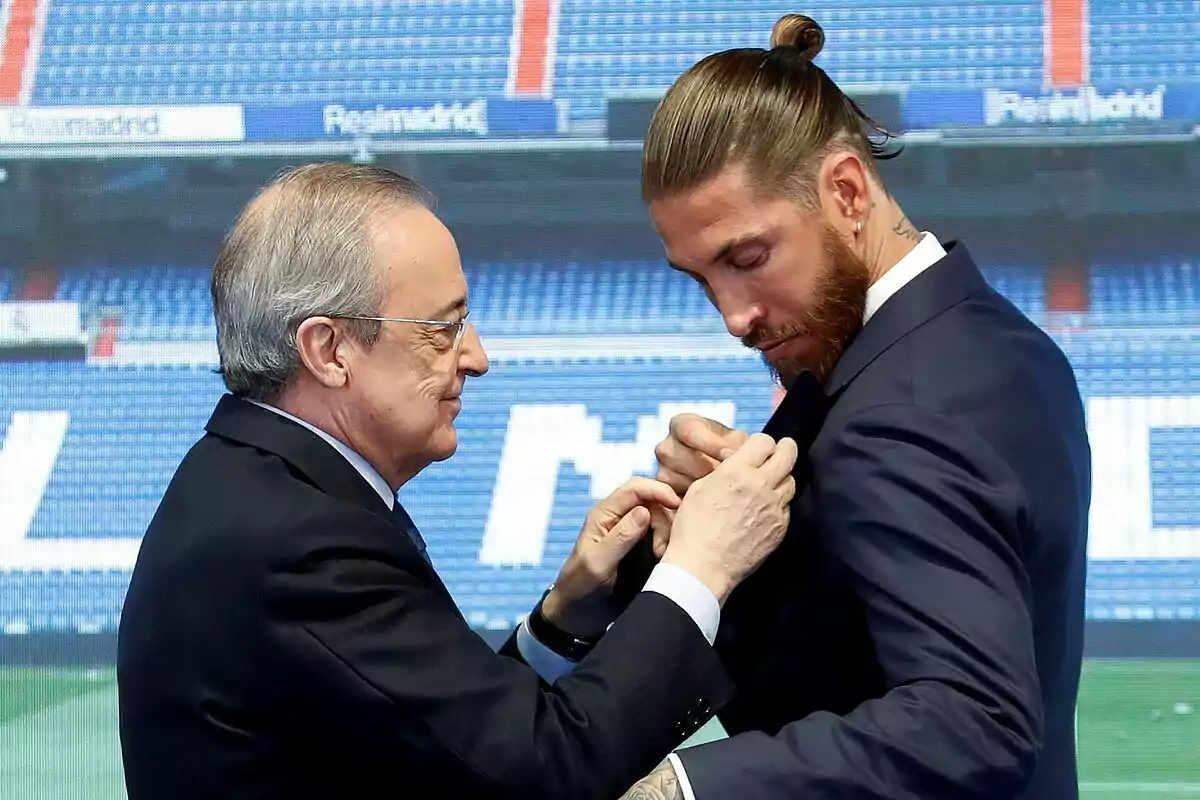 Two men dressed in suits meet in a soccer stadium while one of them adjusts the other’s jacket lapel. Two men dressed in suits meet in a soccer stadium while one of them adjusts the other’s jacket lapel.
