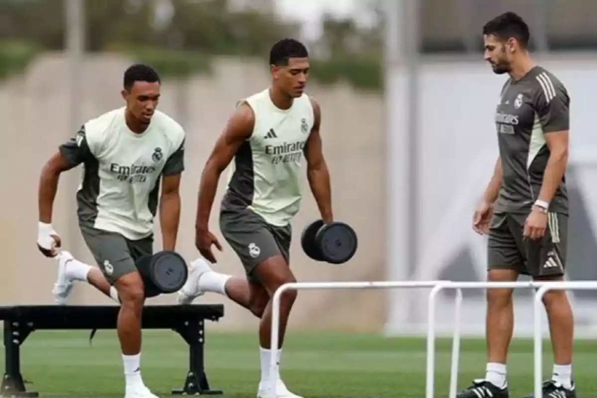 Dos futbolistas entrenan con pesas en un campo mientras un entrenador los observa Dos futbolistas entrenan con pesas en un campo mientras un entrenador los observa