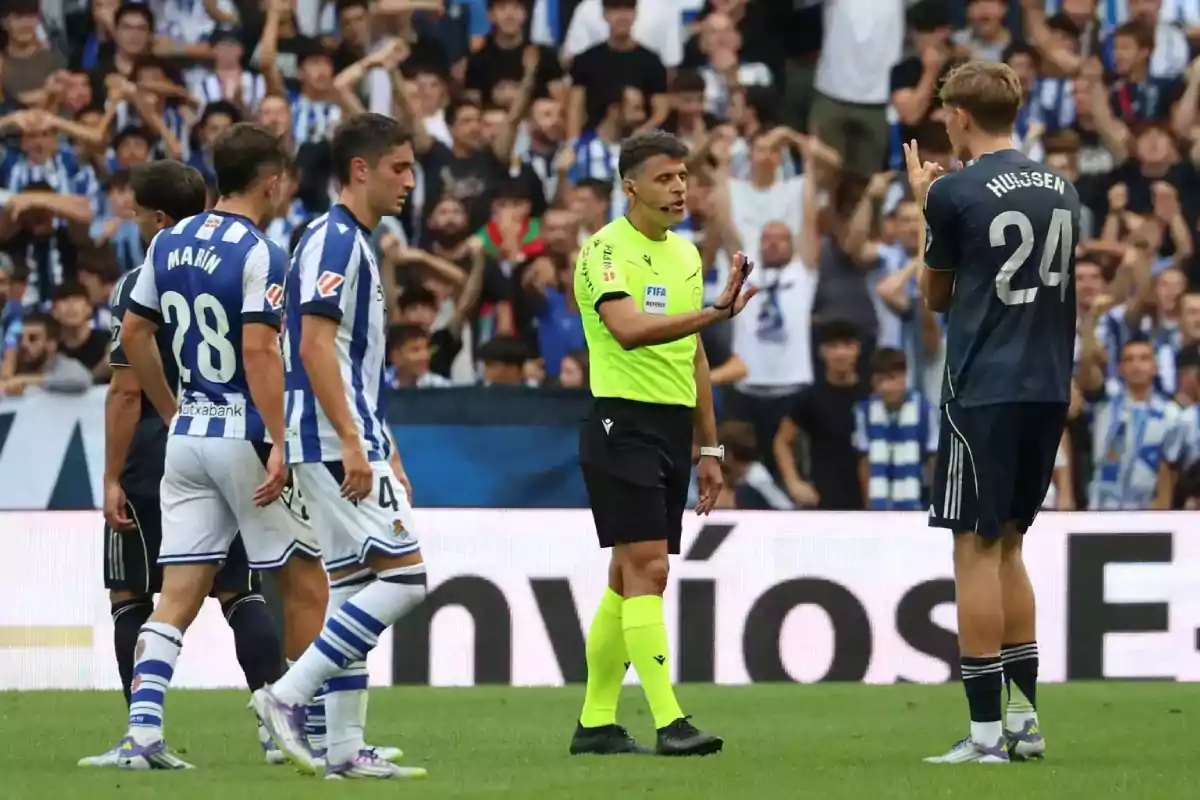 Un árbitro de fútbol detiene el juego mientras habla con un jugador del equipo visitante, rodeado de jugadores del equipo local y con el público en las gradas al fondo. Un árbitro de fútbol detiene el juego mientras habla con un jugador del equipo visitante, rodeado de jugadores del equipo local y con el público en las gradas al fondo.