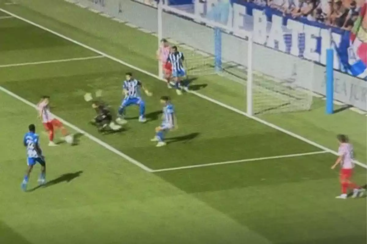 A player in a red uniform tries to score a goal while the goalkeeper and several defenders in blue and white uniforms try to stop him near the goal. A player in a red uniform tries to score a goal while the goalkeeper and several defenders in blue and white uniforms try to stop him near the goal.
