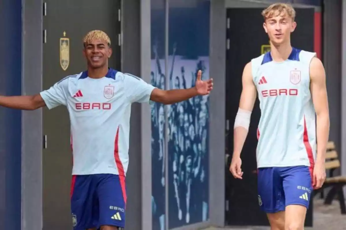 Dos jóvenes futbolistas con uniforme de entrenamiento claro y pantalones cortos azules caminan sonrientes al aire libre frente a un edificio deportivo Dos jóvenes futbolistas con uniforme de entrenamiento claro y pantalones cortos azules caminan sonrientes al aire libre frente a un edificio deportivo