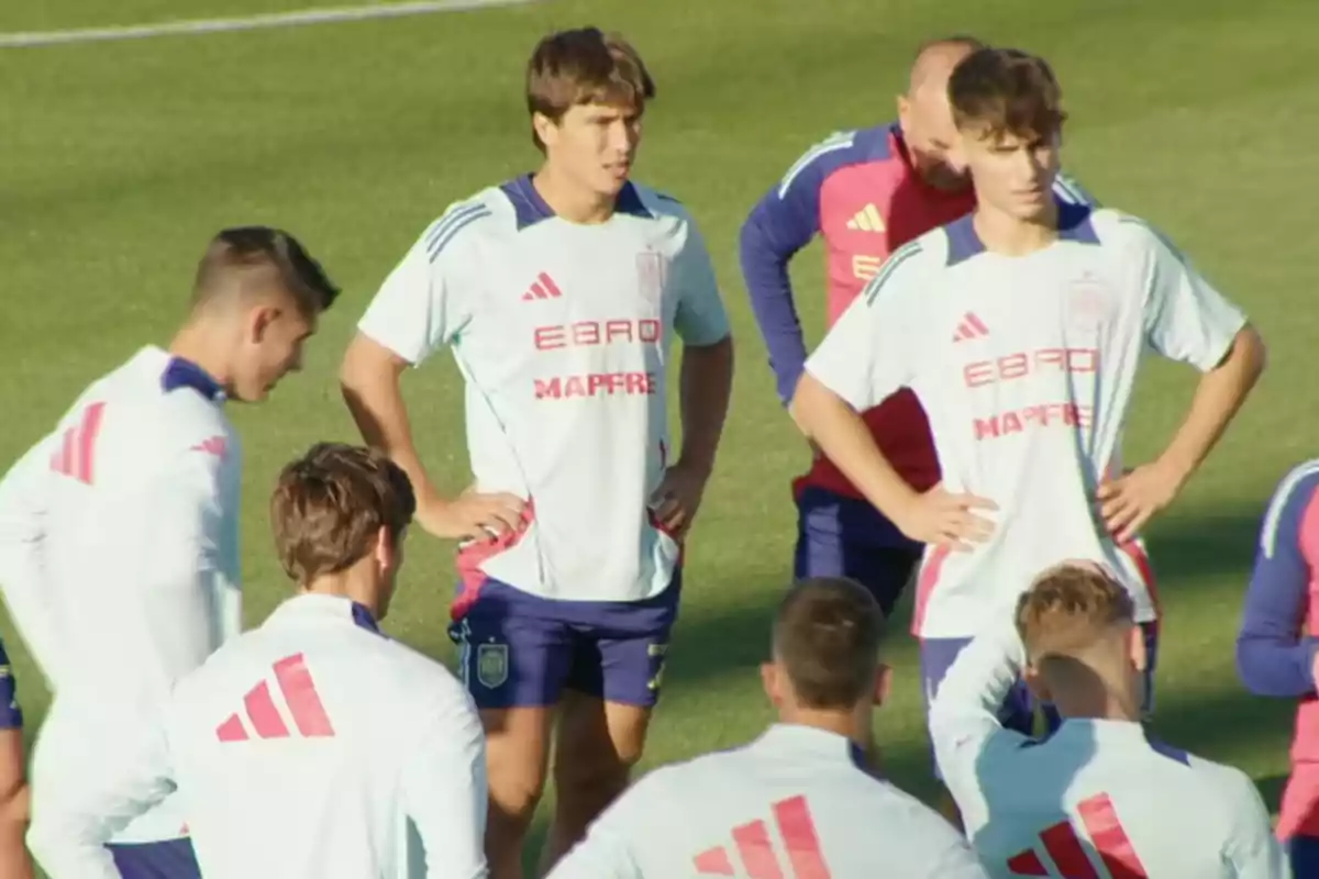 Soccer players gathered on the field during a training session wearing light-colored uniforms with red accents. Soccer players gathered on the field during a training session wearing light-colored uniforms with red accents.
