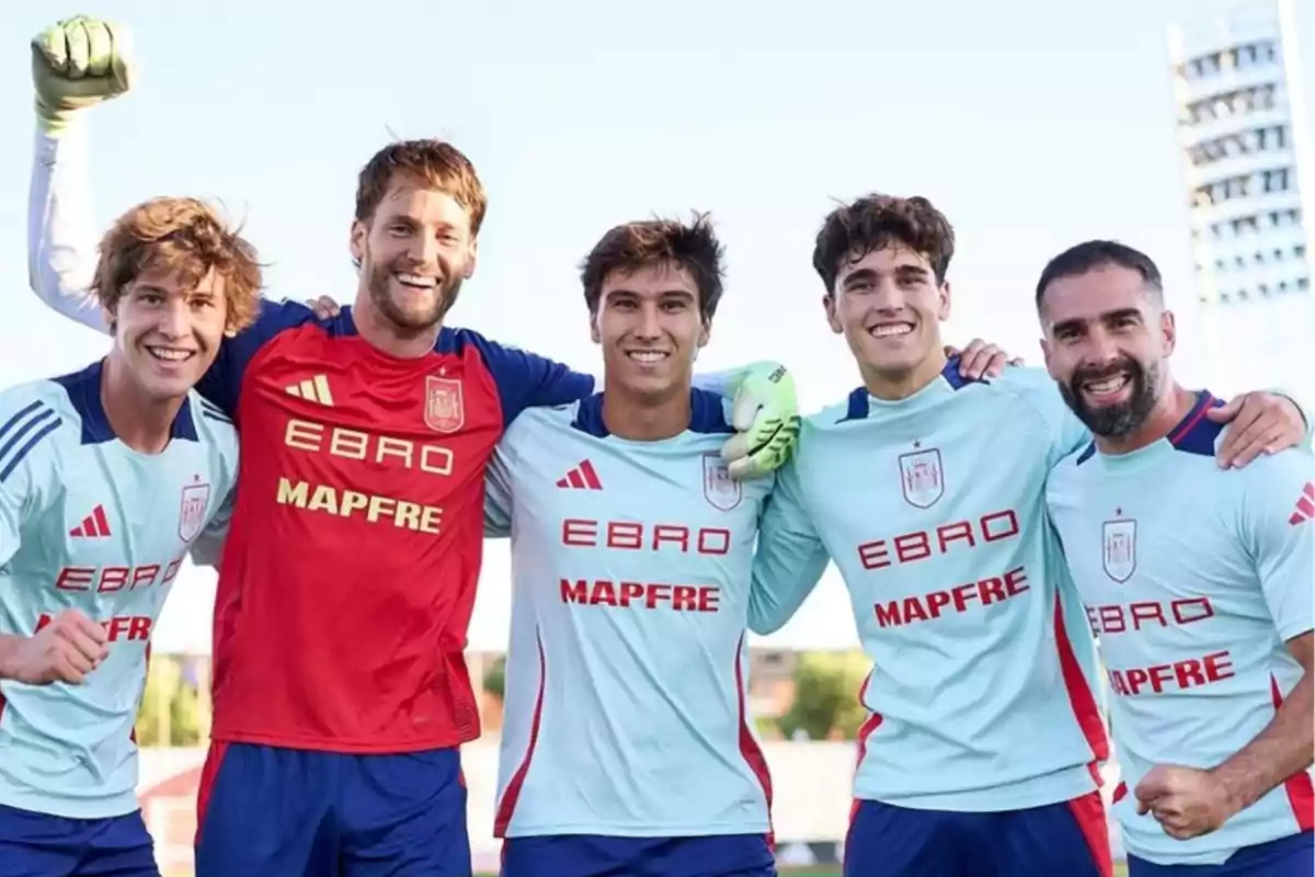 Five players smile and pose with their arms around each other on the field, wearing light blue uniforms and one red, showing camaraderie and joy.