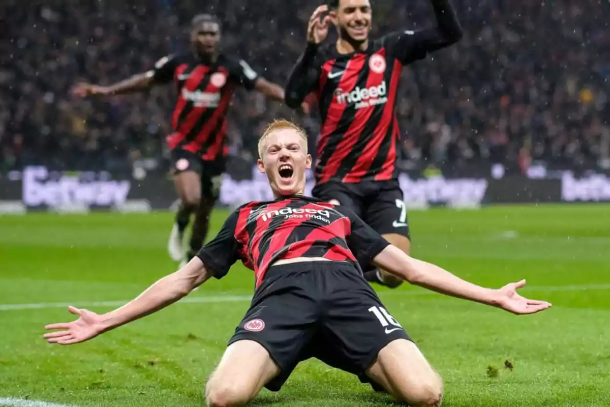 Futbolista celebrando un gol deslizándose de rodillas en el césped mientras sus compañeros corren hacia él con alegría Futbolista celebrando un gol deslizándose de rodillas en el césped mientras sus compañeros corren hacia él con alegría