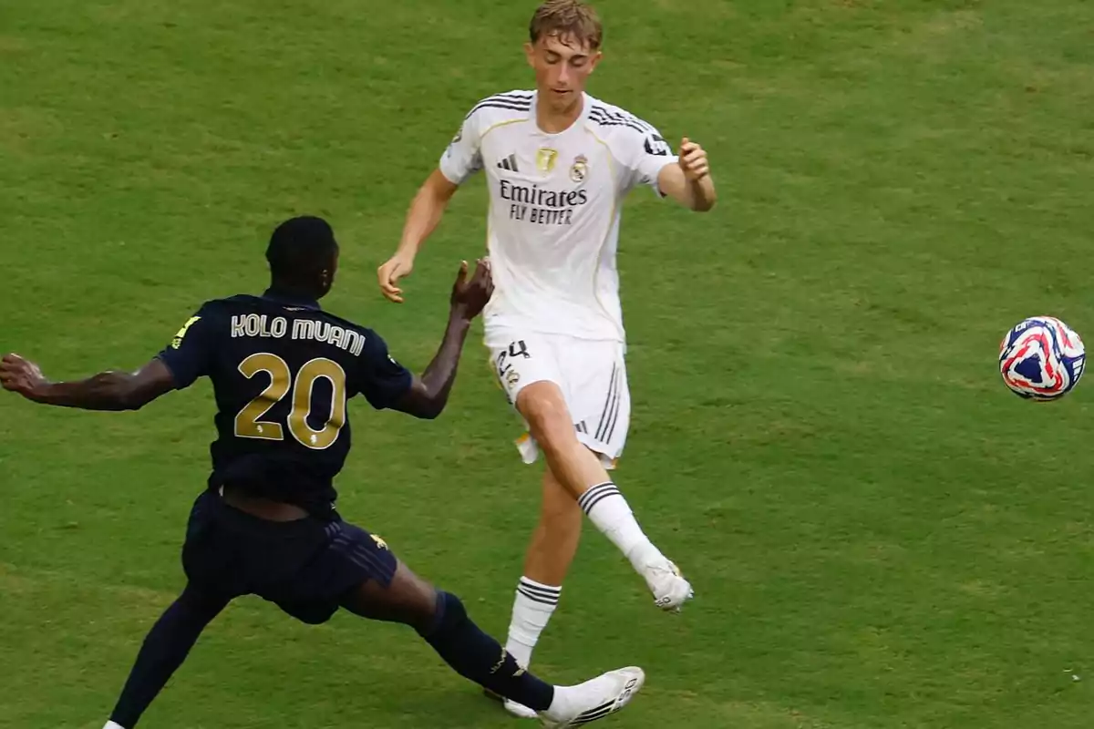 Dos futbolistas disputan el balón en un partido sobre césped uno con uniforme blanco y otro con uniforme oscuro Dos futbolistas disputan el balón en un partido sobre césped uno con uniforme blanco y otro con uniforme oscuro