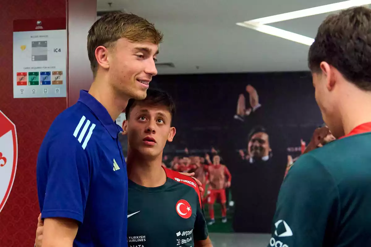 Three young players talk and smile in a stadium hallway; one of them is wearing a blue jersey and the other two are wearing Türkiye national team jerseys.