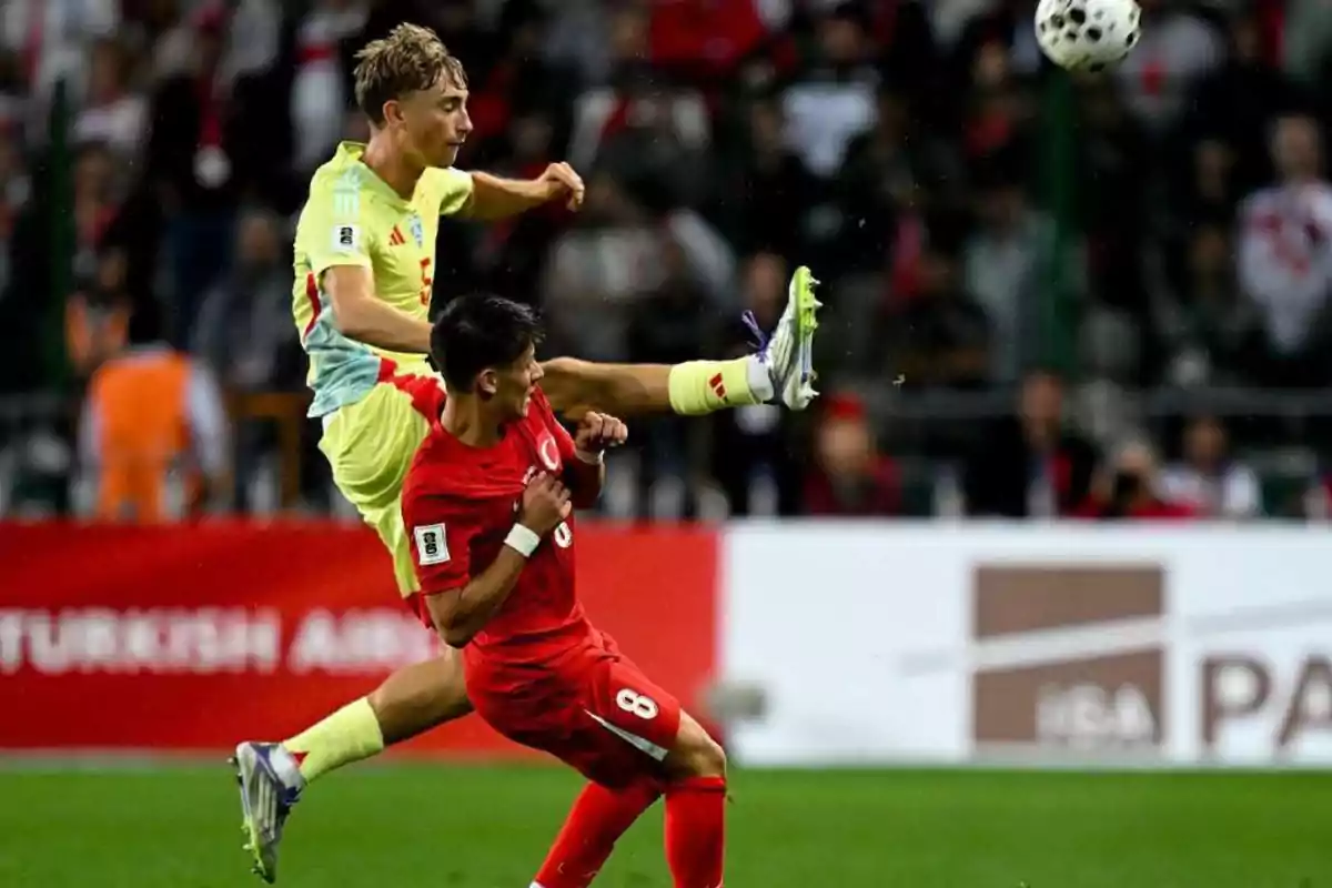 Jugador con uniforme amarillo salta y extiende la pierna mientras otro jugador con uniforme rojo se agacha para evitar el contacto durante un partido de fútbol Jugador con uniforme amarillo salta y extiende la pierna mientras otro jugador con uniforme rojo se agacha para evitar el contacto durante un partido de fútbol