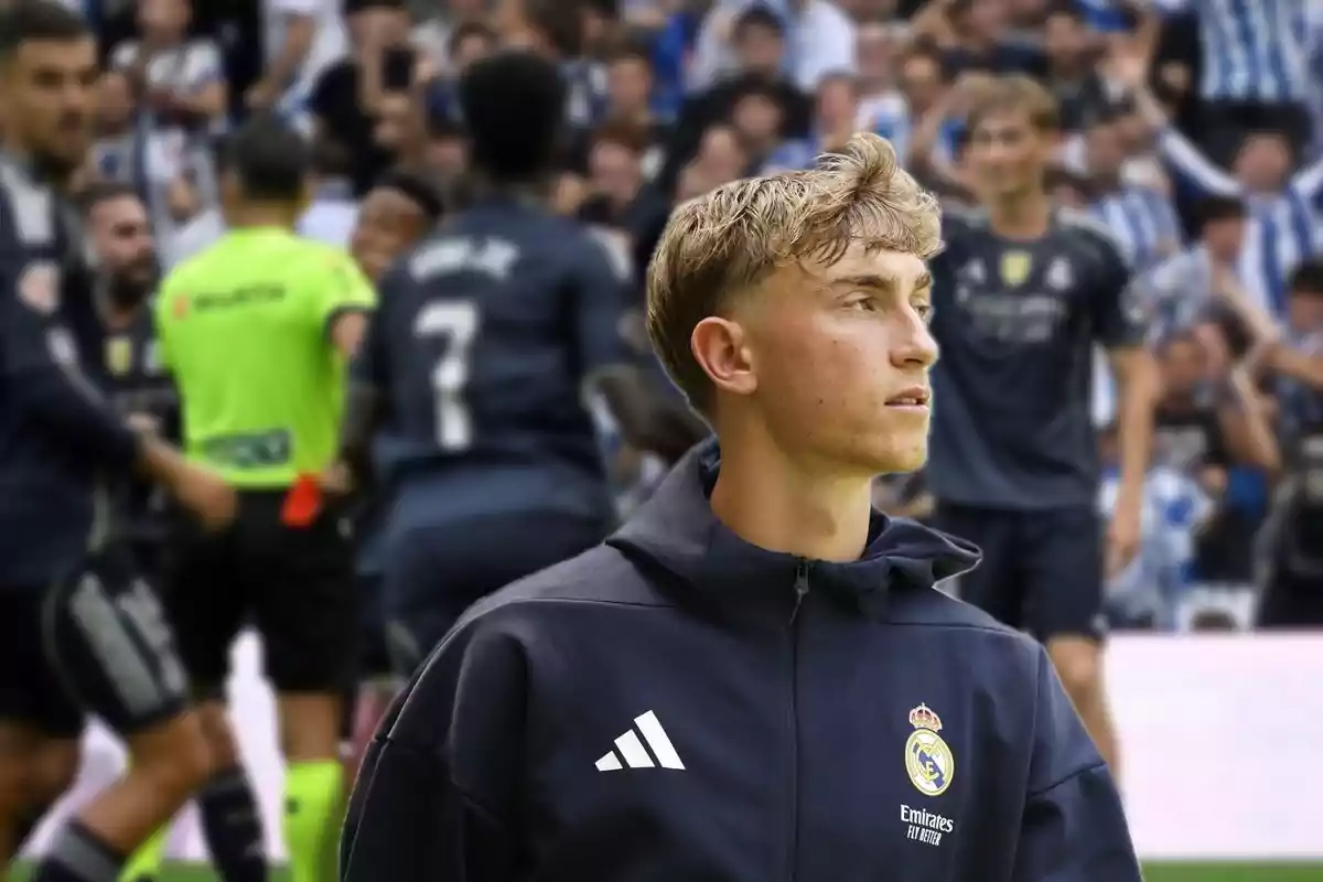 Joven futbolista del Real Madrid con chaqueta del equipo en primer plano mientras de fondo se observa a otros jugadores y un árbitro en un estadio lleno de aficionados